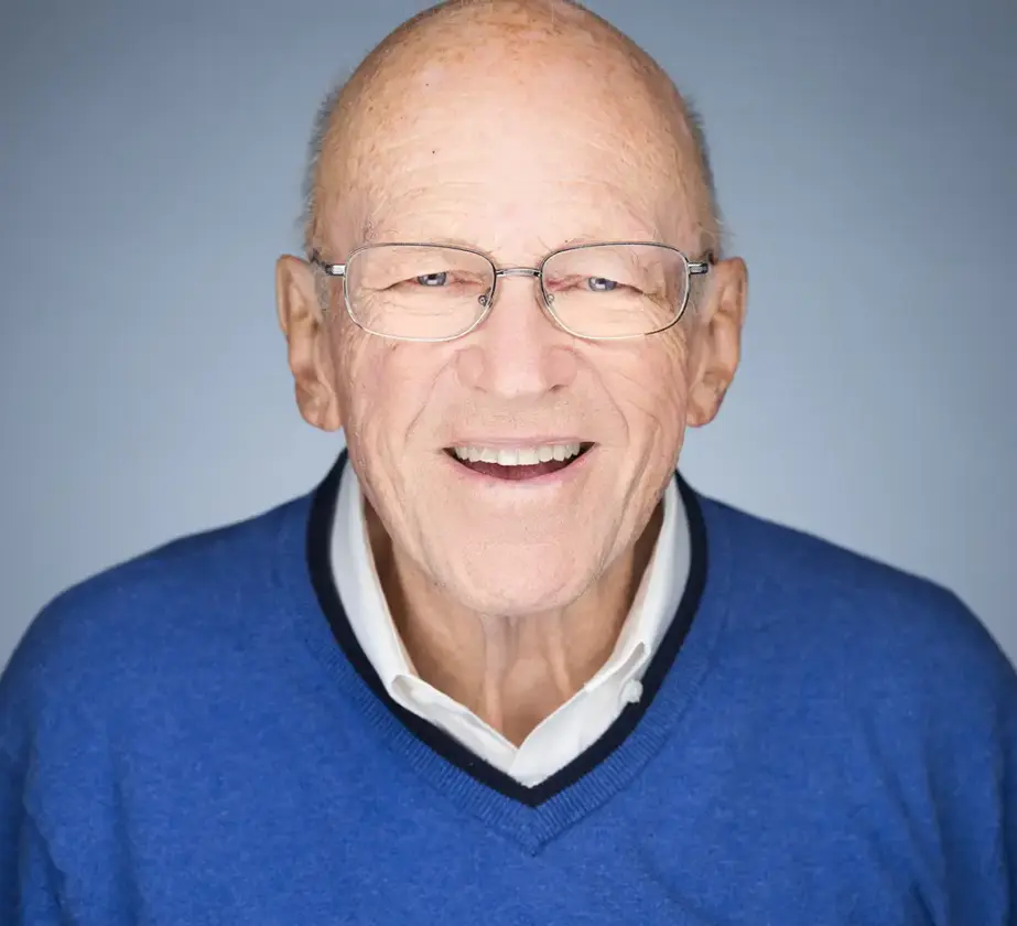 Ken Blanchard, ein älterer Mann mit Brille und Glatze, lächelt warmherzig. Er trägt einen blauen Pullover über einem weißen Hemd mit Kragen und steht vor einem schlichten hellblauen Hintergrund.