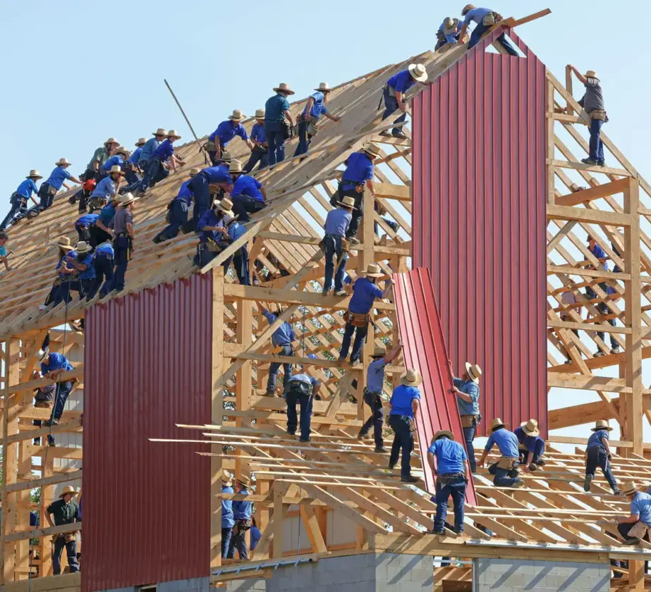 Dutzende von Menschen arbeiten gemeinsam am Holzgerüst einer großen Scheune und montieren unter strahlend blauem Himmel rote Metallpaneele auf dem Dach und an den Wänden, wobei die psychologische Sicherheit am Arbeitsplatz durch die effiziente Zusammenarbeit gefördert wird.