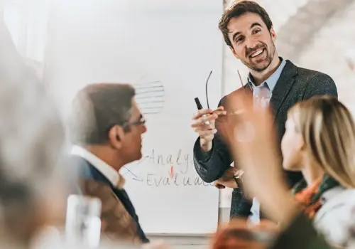 Ein Mann in Businesskleidung lächelt und gestikuliert, während er vor einem Flipchart eine Sitzung zum Thema Pyramidales Präsentieren leitet. Mehrere Kollegen sitzen um einen Tisch und diskutieren. Die Atmosphäre wirkt positiv und kooperativ.