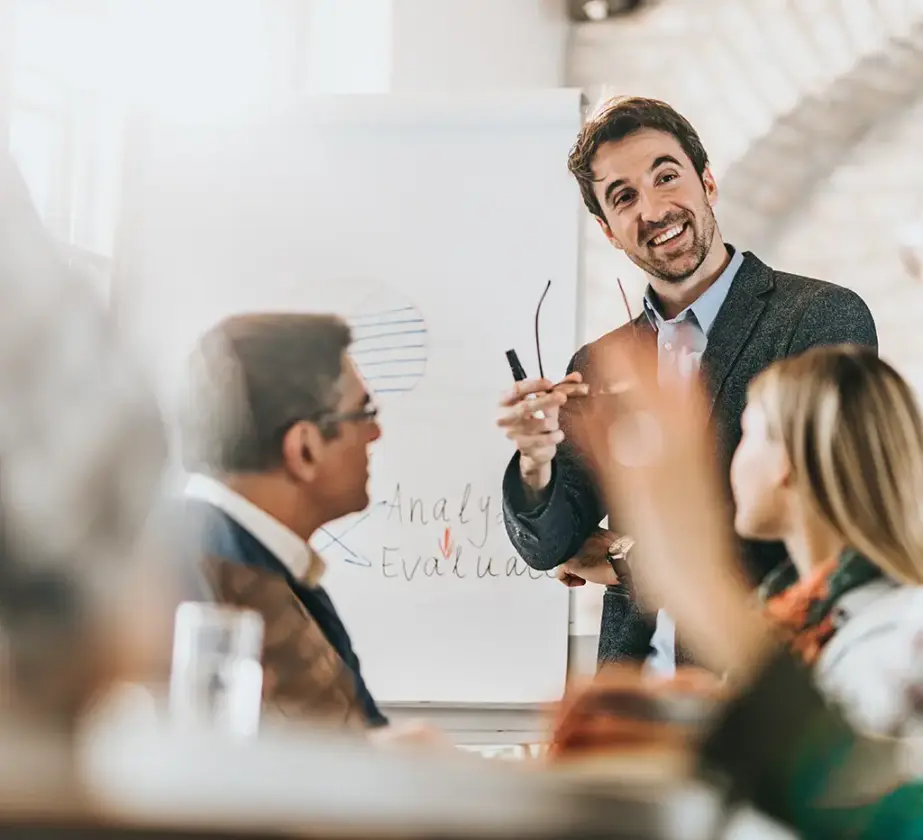 A man in a suit smiles and gestures while presenting learning myths in continuing education to colleagues in a bright office. He stands before a flipchart with graphs and notes, engaging with seated team members.