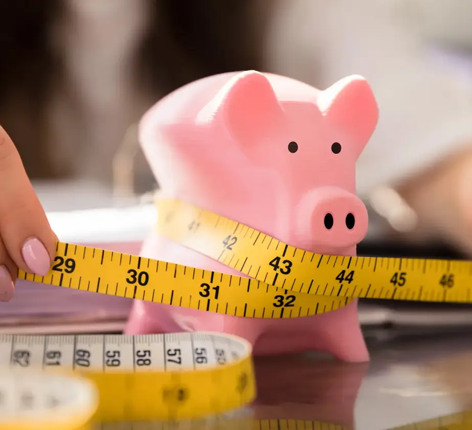Eine Person wickelt ein gelbes Maßband um ein rosa Sparschwein auf einem Schreibtisch und symbolisiert damit Budgetierung, finanzielle Ersparnisse und die Bedeutung der Weiterbildung in Krisenzeiten.