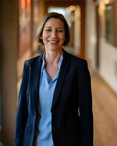 A woman with short brown hair wearing a navy blazer and light blue blouse smiles while standing in a well-lit hallway with artwork on the walls.