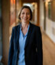 Eine Frau mit kurzen braunen Haaren, einer blauen Bluse und einem dunklen Blazer steht lächelnd in einem gut beleuchteten Flur mit Kunstwerken und Fotos an den Wänden.