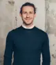 Vincent Aretz. ein Mann mit langen, zurückgebundenen Haaren und einem kurzen Bart lächelt in die Kamera. Er trägt einen dunklen Pullover über einem weißen Kragenhemd und steht vor einer strukturierten, verwitterten Wand.