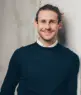 Vincent Aretz, ein Mann mit hellem Bart und langen, nach hinten gebundenen Haaren lehnt lächelnd an einer hellen, strukturierten Wand. Er trägt einen dunklen Pullover über einem weißen Hemd mit Kragen.