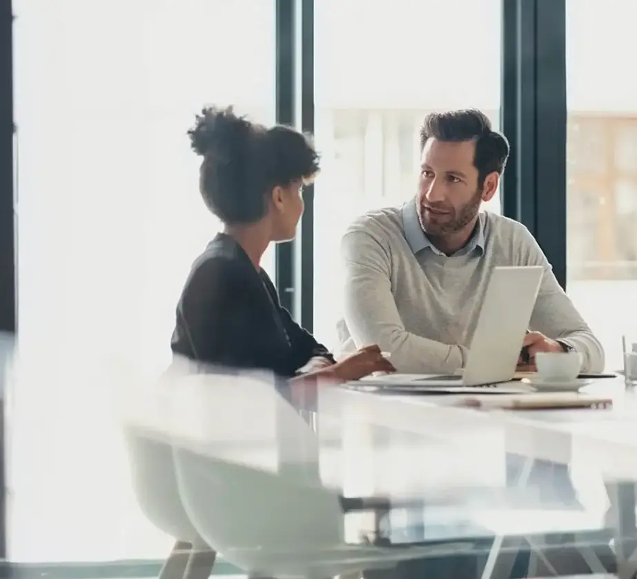 Zwei Personen sitzen an einem Schreibtisch in einem hellen Büro und unterhalten sich. Eine Person hat einen Laptop geöffnet, auf dem Tisch liegen Papiere und eine Tasse. Große Fenster lassen Tageslicht herein.