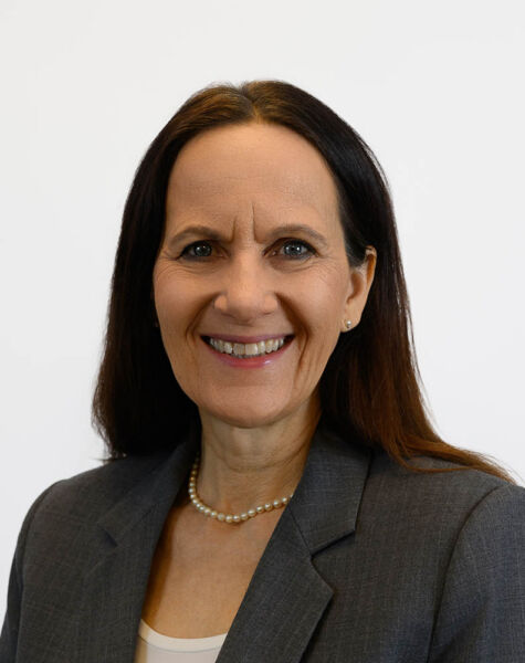 Gesa Schubert, eine Frau mit langen braunen Haaren, die einen grauen Blazer, eine Perlenkette und ein helles Top trägt, lächelt vor einem schlichten weißen Hintergrund.