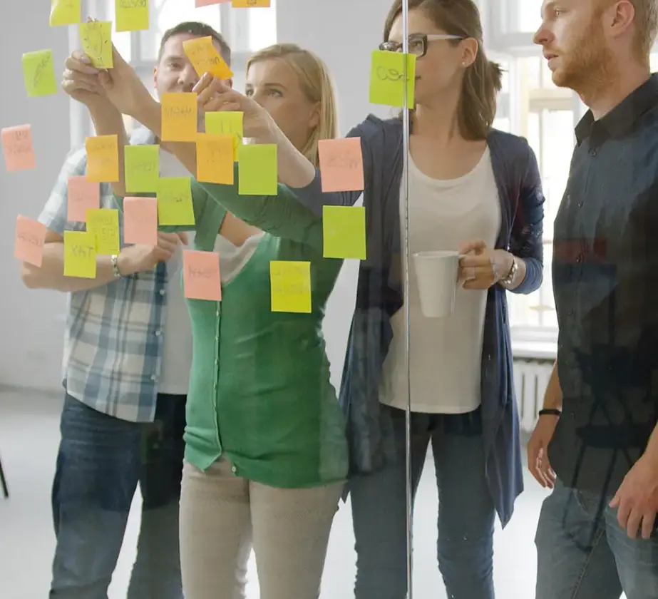 Gruppe von Meetingteilnehmenden clustert PostIts an einer Glaswand im Besprechungsraum