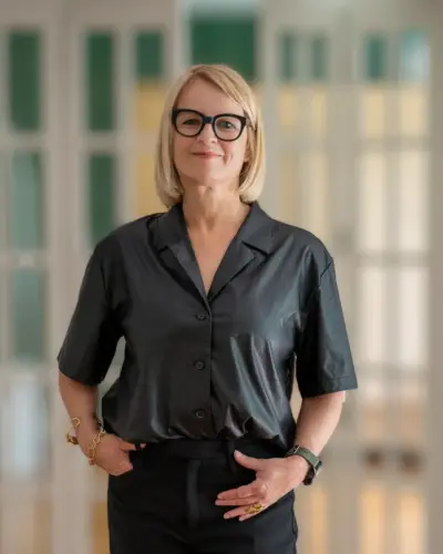 A woman with short blonde hair and glasses wears a black short-sleeve button-up shirt and black pants, standing indoors in front of glass-paneled doors, smiling confidently.