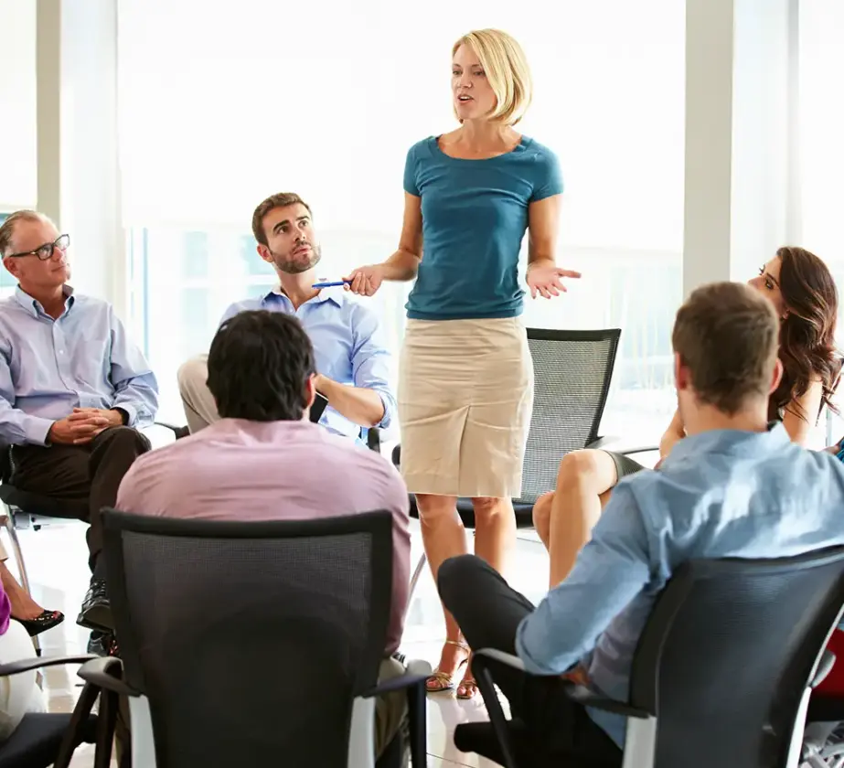 Eine Frau steht in einem Büro und spricht zu einer Gruppe von Menschen, die im Kreis sitzen. Sie schlägt eine Besprechung oder eine Gruppendiskussion vor. Die Gruppe hört aufmerksam zu, einige blicken in die Richtung der Sprecherin. Der Raum ist hell und hat große Fenster.
