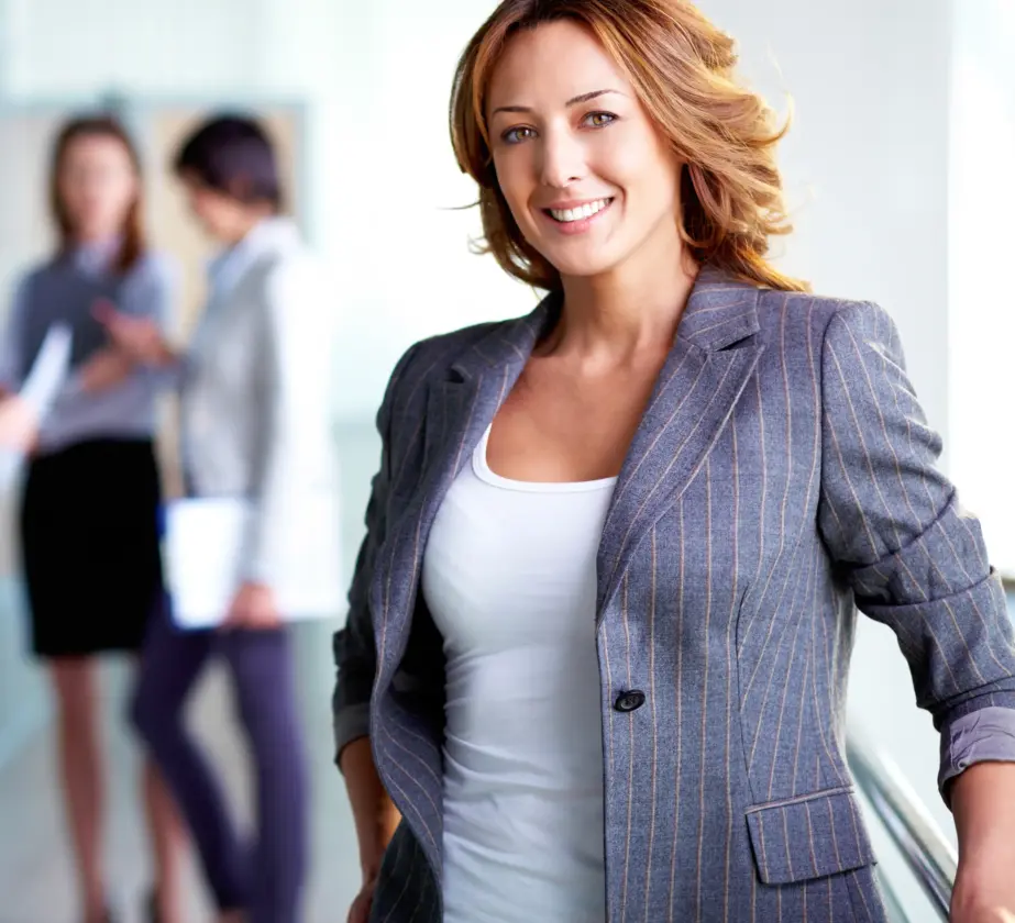 Eine selbstbewusste Frau in einem Nadelstreifenblazer und einem weißen Oberteil lächelt in einem hellen Büroflur in die Kamera, während sich im unscharfen Hintergrund zwei Kollegen unterhalten.
