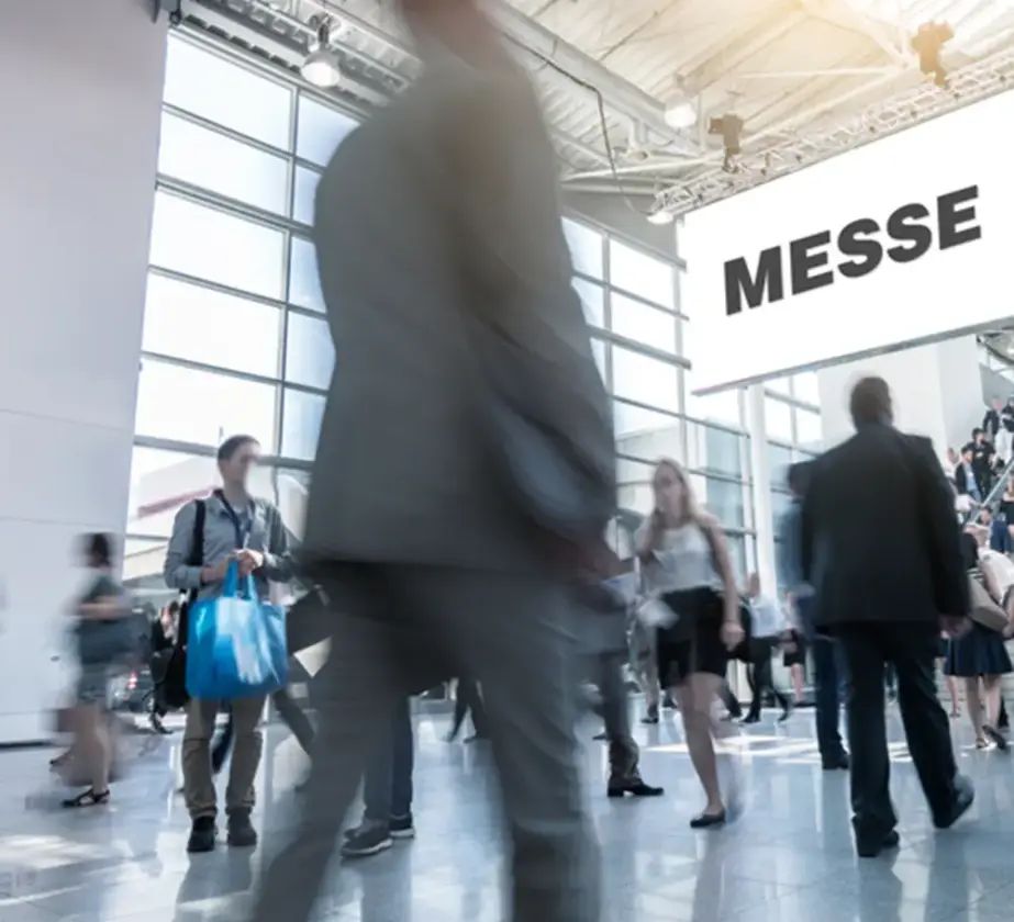 Menschen gehen durch eine helle, moderne Messehalle mit hohen Fenstern. Ein großes Schild darüber MESSE suggeriert eine Messe oder ein Messetraining. Die Szene ist voller verschwommener Bewegungen und lebhafter Aktivität.