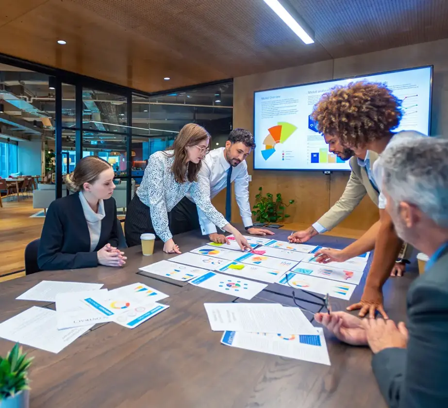 Fünf Geschäftsleute versammeln sich um einen mit Diagrammen und Dokumenten bedeckten Konferenztisch und diskutieren aktiv Daten und Teamführungsstrategien. Im Hintergrund des modernen Büros ist ein großer Bildschirm mit bunten Grafiken zu sehen.