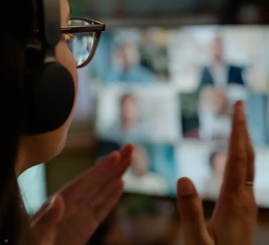 Eine Person mit Brille und Kopfhörern gestikuliert während einer virtuellen Präsentation während eines Videoanrufs, wobei auf einem Computerbildschirm im Hintergrund mehrere verschwommene Gesichter sichtbar sind.