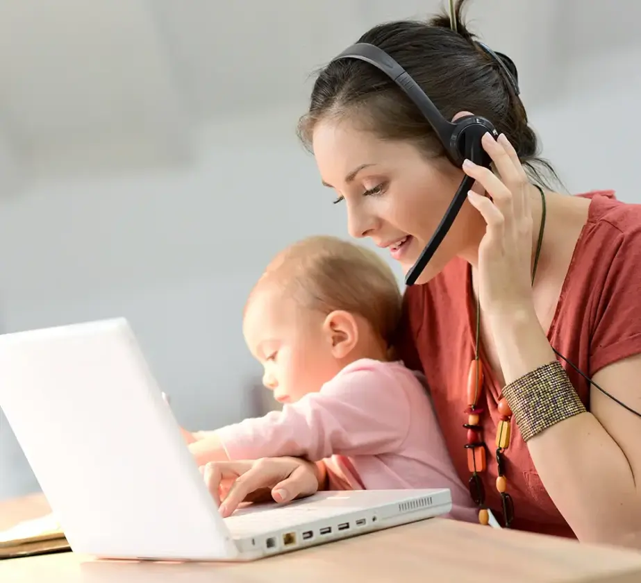 Eine Frau mit Headset arbeitet an einem Laptop, während sie ein Baby auf dem Schoß hält. Sie ist multitaskingfähig und konzentriert, während sie versucht, ihr Zeitmanagement im Arbeitsalltag zu verbessern. Das Baby greift neugierig nach der Tastatur.