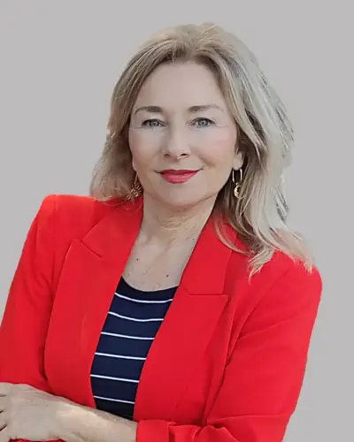 A woman with long blonde hair wearing a bright red blazer and a black and white striped top stands against a plain gray background, smiling confidently.