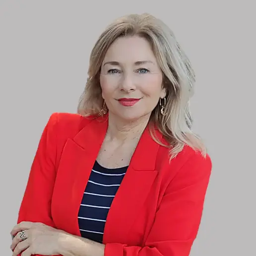 A woman with long blonde hair wearing a bright red blazer and a black and white striped top stands against a plain gray background, smiling confidently.