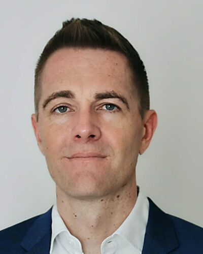 A man with short brown hair, light skin, and blue eyes wearing a dark blue suit jacket and white collared shirt, standing against a plain light background.