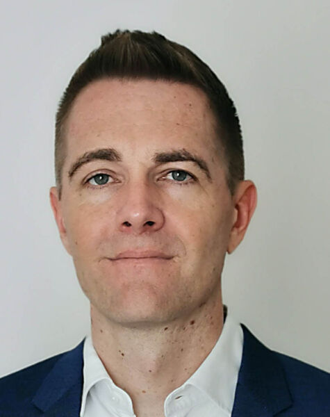 A man with short brown hair, light skin, and blue eyes wearing a dark blue suit jacket and white collared shirt, standing against a plain light background.