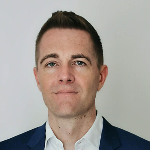 A man with short brown hair, light skin, and blue eyes wearing a dark blue suit jacket and white collared shirt, standing against a plain light background.