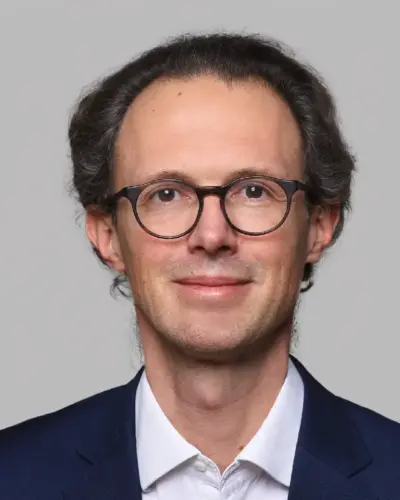 A man with short, wavy dark hair, wearing round black glasses, a white shirt, and a navy blazer, smiles slightly against a plain light gray background.
