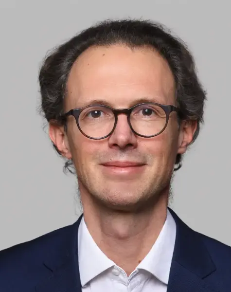 A man with short, wavy dark hair, wearing round black glasses, a white shirt, and a navy blazer, smiles slightly against a plain light gray background.