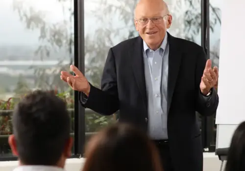 Ken Blanchard steht da und gestikuliert, während er mit einer Gruppe vor ihm sitzender Menschen spricht.