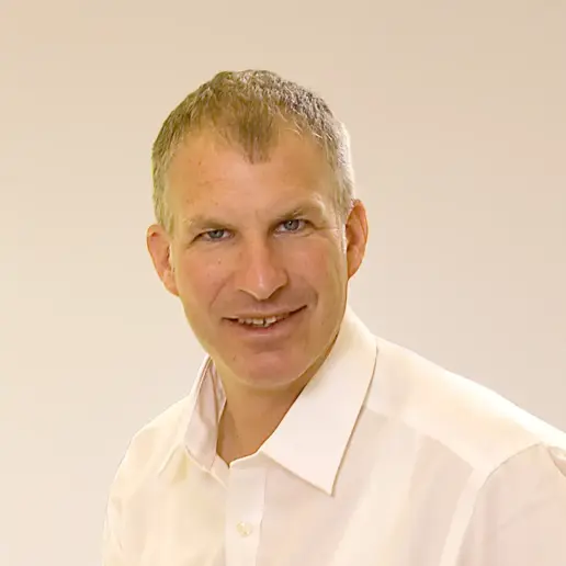 A middle-aged man with short gray hair is smiling and wearing a white collared shirt, posing in front of a plain light-colored background.