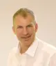 A middle-aged man with short gray hair is smiling and wearing a white collared shirt, posing in front of a plain light-colored background.
