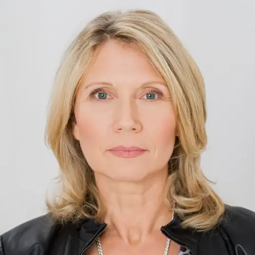 A middle-aged woman with shoulder-length blonde hair and blue eyes looks directly at the camera. She is wearing a black jacket and a light top, with a neutral facial expression against a plain background.