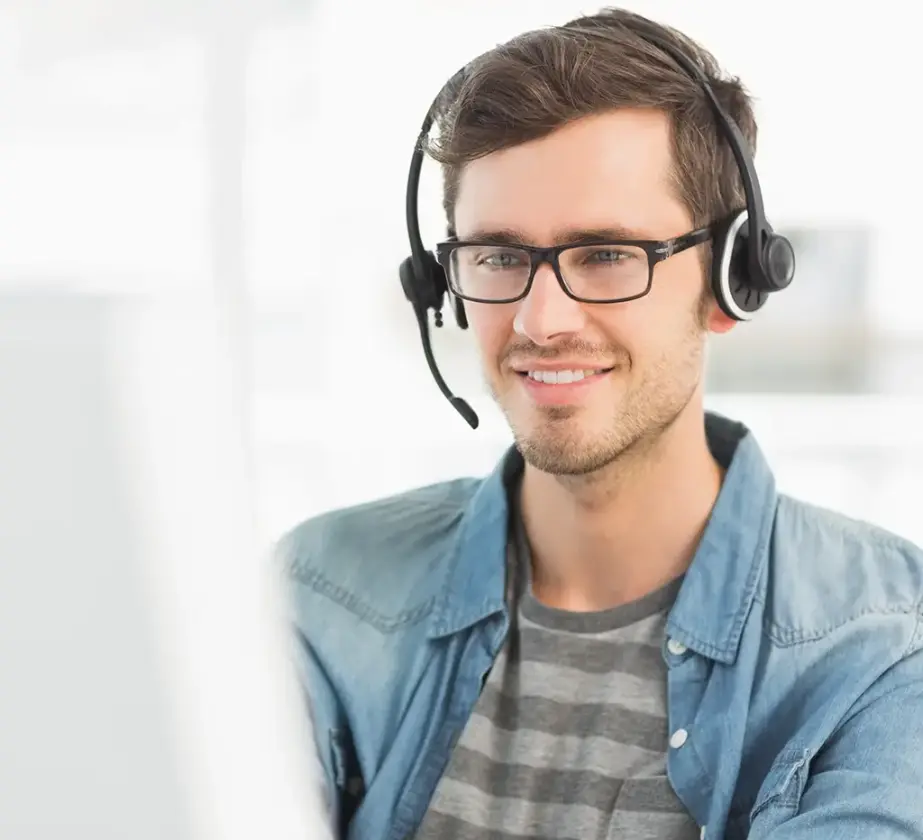 Ein junger Mann mit Brille und Headset lächelt, während er auf einen Computerbildschirm blickt. Er trägt ein lässiges gestreiftes Hemd und eine Jeansjacke und sitzt in einem hellen Büro.