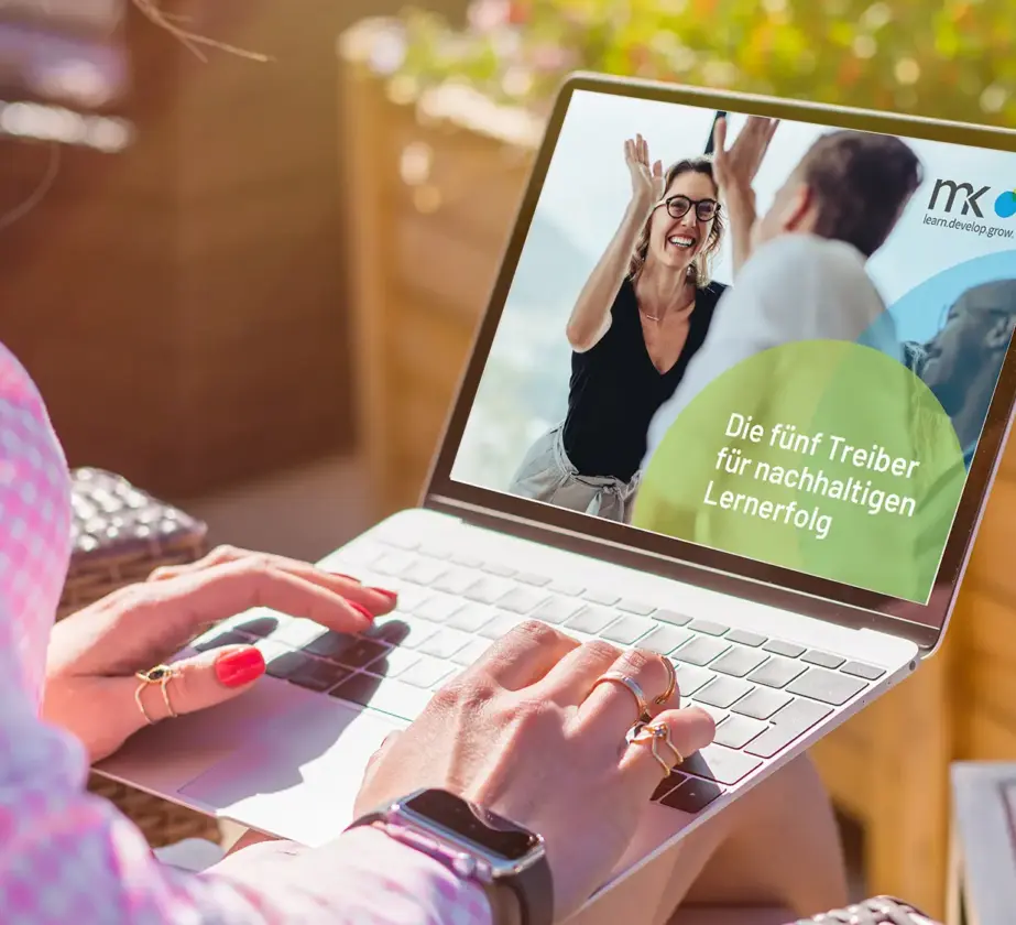 Eine Person, die im Freien einen Laptop benutzt, sieht einen Bildschirm, auf dem eine lächelnde Frau einem Mann ein High Five gibt. Der deutsche Text lautet: „Die fünf Treiber für nachhaltigen Lernerfolg“. Im Hintergrund sind bunte Blumen und Sonnenlicht zu sehen.