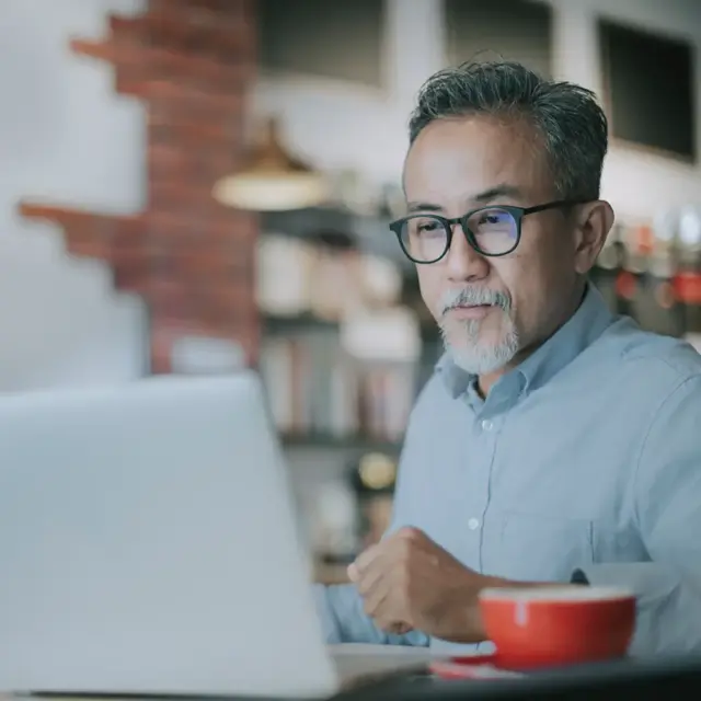 Ein Mann mittleren Alters mit Brille und grauem Haar sitzt an einem Tisch in einem gemütlichen Café und schaut auf seinen Laptop. Vor ihm steht eine rote Kaffeetasse auf dem Tisch, im Hintergrund sind Regale und Lampen verschwommen.