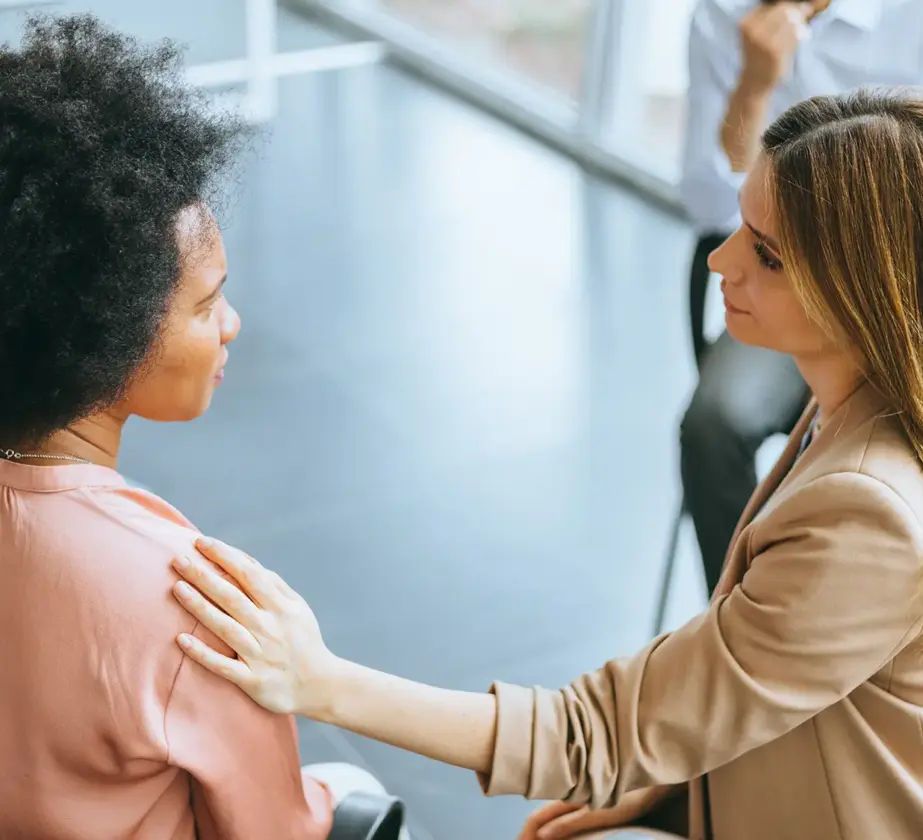 Eine Frau in einem beigen Blazer legt ihre Hand sanft auf die Schulter einer anderen Frau in einem rosa Oberteil und bietet Trost während eines Gruppentreffens in einem hellen, modernen Raum, in dem andere in der Nähe sitzen.