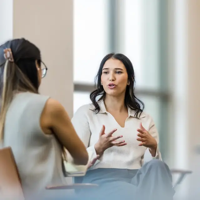 Zwei Frauen sitzen sich im Gespräch gegenüber und nehmen am Kommunikationscoaching teil. Die eine gestikuliert mit den Händen, die andere hört aufmerksam zu. Beide sitzen in einem hellen Raum mit großen Fenstern.