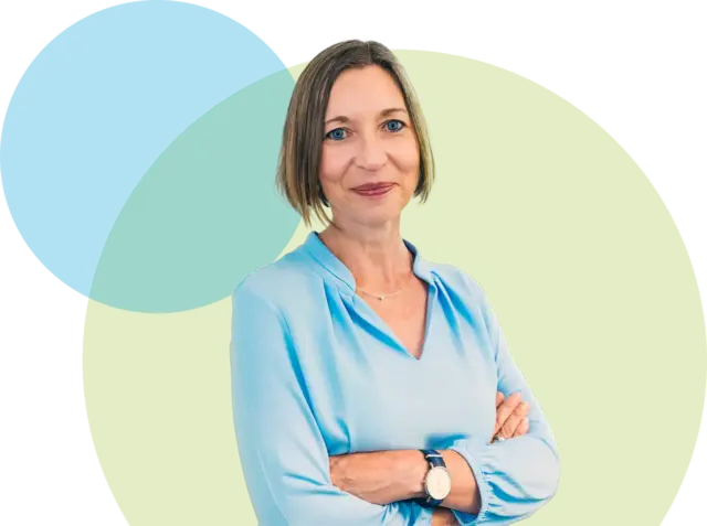 Eine Frau mit kurzen hellbraunen Haaren, die eine hellblaue Bluse und eine Armbanduhr trägt, steht mit verschränkten Armen vor sich überlappenden pastellgrünen und blauen Kreisen auf weißem Hintergrund.