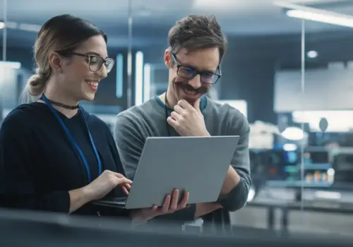 Zwei Personen mit Brille und Ausweis stehen in einem modernen Büro zusammen und lächeln auf einen Laptop-Bildschirm, während sie über Künstliche Intelligenz in der Personalentwicklung diskutieren. Der helle Raum besticht durch eine Glaswand und sichtbare Computerausstattung.