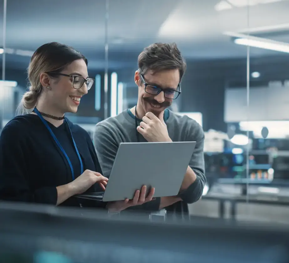Zwei Personen mit Brille und Ausweis stehen in einem modernen Büro zusammen und lächeln auf einen Laptop-Bildschirm, während sie über Künstliche Intelligenz in der Personalentwicklung diskutieren. Der helle Raum besticht durch eine Glaswand und sichtbare Computerausstattung.