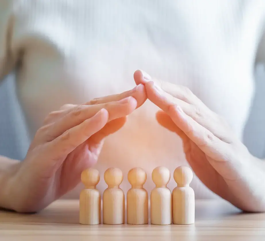Eine Person in einem weißen Hemd formt mit ihren Händen über vier hölzernen menschlichen Figuren auf einem Tisch eine schützende Dachform und symbolisiert damit den Schutz oder die Fürsorge für Menschen oder eine Familie.