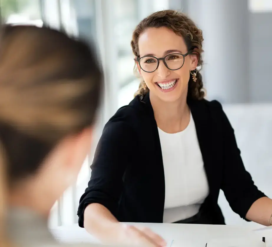 Eine Frau mit lockigem Haar und Brille lächelt, während sie an einem Schreibtisch sitzt und sich mit einer anderen Person, die mit dem Rücken zur Kamera steht, in einer Büroumgebung über Lernformate für betriebliche Weiterbildung unterhält.