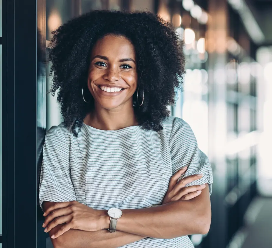 A woman with curly hair, wearing a light striped blouse and watch, stands indoors with her arms crossed, smiling confidently in a modern office hallway with glass walls—reflecting her expertise in personality models in continuing education.
