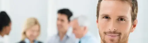 Eine Nahaufnahme eines lächelnden Mannes mit kurzen Haaren und Bart, mit einer verschwommenen Gruppe in Geschäftskleidung, die sich im Hintergrund unterhält - eine Büroszene, die erfolgreiche Trainings- und Coachingprojekte widerspiegelt.