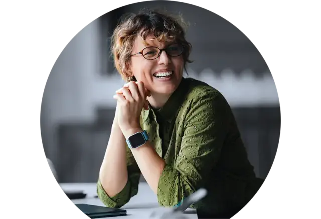 Eine Person mit kurzen Locken, Brille und einem grünen Hemd sitzt an einem Schreibtisch und lächelt. Sie trägt eine Smartwatch und stützt die Hände am Kinn ab; sie wirkt fröhlich und konzentriert.