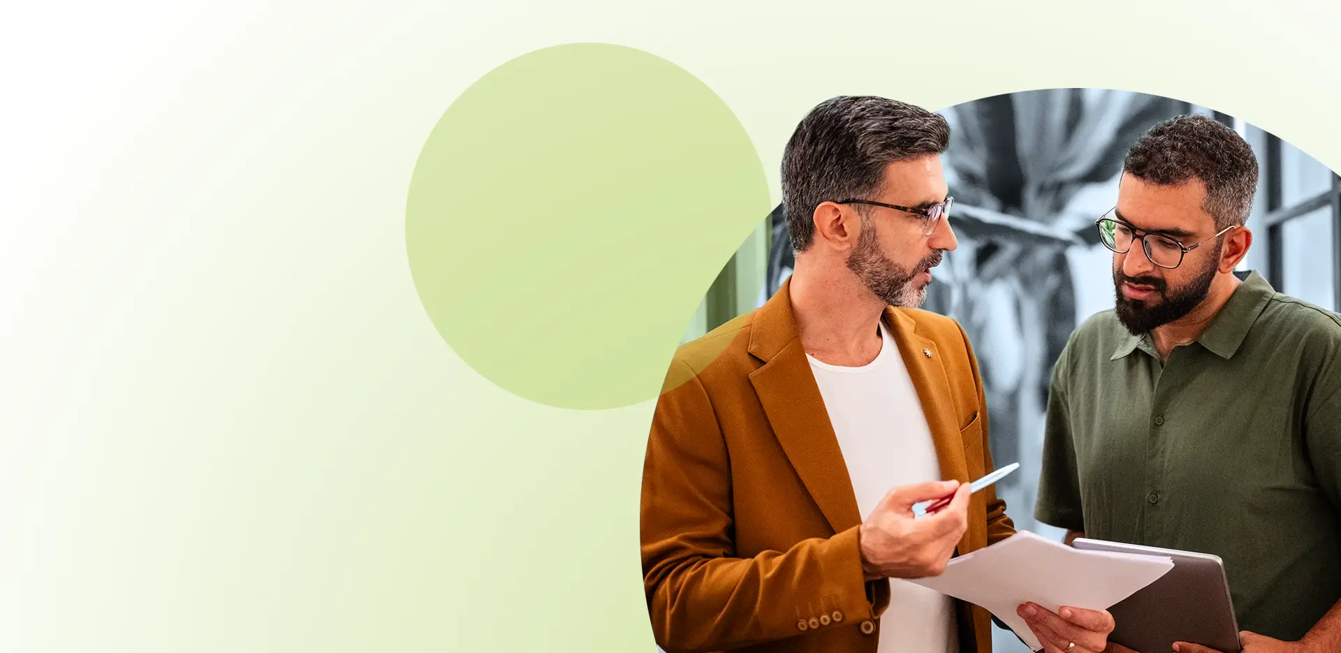 Zwei Männer stehen da und besprechen Unterlagen; der eine hält einen Stift in der Hand, der andere ein Tablet, während sie sich im Rahmen eines Konfliktmanagement-Seminars für Unternehmen in ein aufschlussreiches Gespräch vertiefen. Der Hintergrund ist hell und mit abstrakten grünen Kreisen gestaltet.