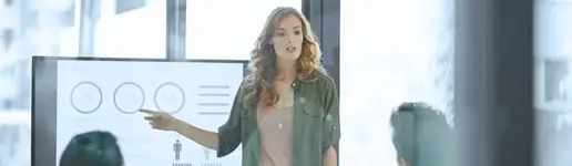 Eine Frau steht vor einem Bildschirm, auf dem Diagramme und Grafiken zu sehen sind, und zeigt auf die Daten, während sie mit zwei sitzenden Personen in einem hellen, modernen Büro spricht – ein idealer Rahmen für erfolgreiche Trainings- und Coachingprojekte.