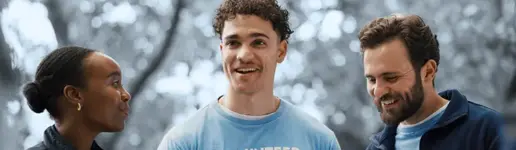 Drei Personen in blauen „VOLUNTEER“-T-Shirts stehen gemeinsam im Freien, lächeln und unterhalten sich. Der Hintergrund mit den Bäumen ist unscharf, wodurch der freundliche Umgang der Gruppe besonders hervorgehoben wird – ein perfektes Beispiel für Zusammenarbeit in erfolgreichen Trainings- und Coaching-Projekten.