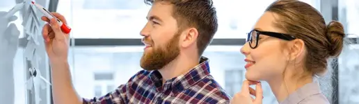 Ein bärtiger Mann schreibt mit einem roten Stift auf eine Weißwandtafel, während eine Frau mit Brille lächelnd zusieht; beide wirken engagiert und kooperativ – eine ideale Szene, die erfolgreiche Trainings- und Coachingprojekte widerspiegelt.