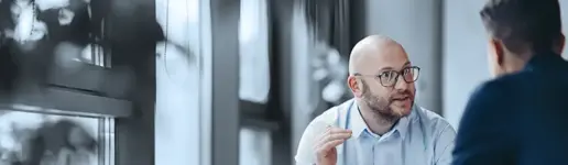 Zwei Männer sitzen an einem Tisch in der Nähe großer Fenster und führen ein ernstes Gespräch. Der eine Mann, der eine Brille und ein hellblaues Hemd trägt, gestikuliert mit den Händen, während er mit dem anderen spricht, der von hinten zu sehen ist. Ihre konzentrierte Diskussion spiegelt die Atmosphäre erfolgreicher Trainings- und Coachingprojekte wider.