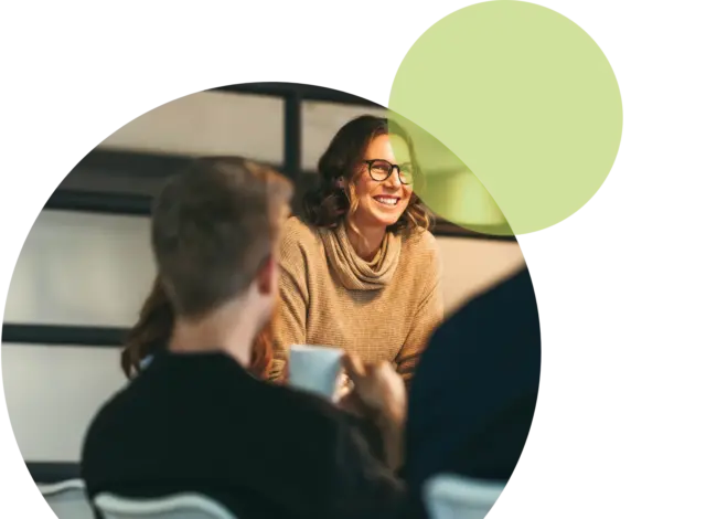 Eine Frau mit Brille sitzt mit zwei weiteren Personen an einem Tisch und nimmt an einer Teamentwicklung für Unternehmen teil. Eine der Personen hält einen weißen Becher in der Hand. Im Hintergrund sind eine moderne Einrichtung sowie abstrakte grüne und weiße Kreisformen zu sehen.