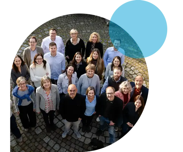 Eine bunt gemischte Gruppe von 20 Personen – Männer und Frauen unterschiedlichen Alters – steht im Kreis und blickt nach oben, was die Werte und Haltung von „mensch & kommunikation“ widerspiegelt. Oben rechts im Bild ist ein blauer Kreis zu sehen.
